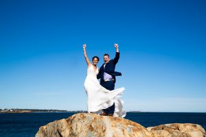A bride and groom standing on a huge rock formation with the ocean behind them . Robin and Tim on the rocks at bass rocks Gloucester MA!